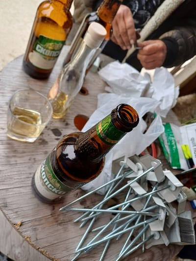 The image depicts a close-up, high-angle view of a cluttered wooden table surface, located in Prdejci, North Macedonia.

On the table, several objects are visible:
*   A clear glass tumbler containing a light amber liquid, positioned in the lower-left.
*   An open amber glass bottle with a green and white label prominently featuring the text "CKONCKO" (Skopsko), positioned diagonally from the lower-left.
*   Two other amber glass bottles, one partially obscured behind the tumbler and another visible in the background, suggesting additional beverages.
*   An empty clear glass bottle with a light-colored stopper, standing upright.
*   A significant pile of silver-colored threaded rods, each fitted with a white plastic or metal bracket, resembling construction anchors or specialized fasteners. Some of these fasteners feature small brush-like components.
*   Crumpled pieces of white paper or plastic material are scattered among the items.
*   In the upper right, a pair of hands with dark skin and a visibly calloused texture are engaged in manipulating a small, thin, metallic object. The individual is wearing a dark jacket with a distinct, textured, possibly sheepskin-lined collar.
*   Additional items, partially visible and out of focus in the background, include what appears to be a red tool (possibly pliers), a green and white packaged item, and yellow and black writing instruments.

The scene suggests an informal gathering or break, potentially associated with construction or manual labor, with the presence of both beverages and work-related materials.