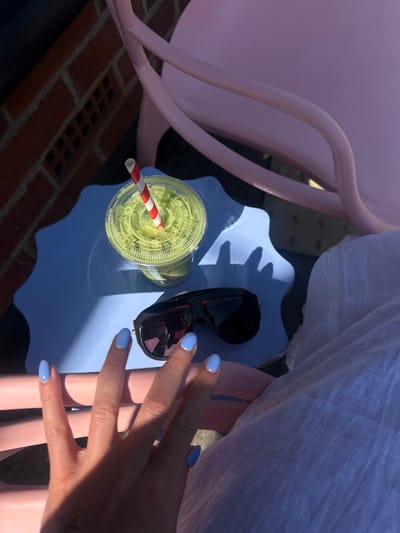 This media file captures a top-down, slightly angled view of an outdoor setting on a sunny day. The primary subjects are a person's hand, a beverage, and sunglasses.
A hand, featuring light blue nail polish, is visible on the right side, with its shadow cast across a light blue, scalloped-edge table. Resting on this table, next to the hand, are a pair of black sunglasses with dark lenses. To the left of the sunglasses is a clear plastic cup filled with a bright green beverage, likely matcha or a smoothie, and a red and white striped straw. The transparent plastic lid of the cup has concentric circular patterns, and partial text "TABLE ONE COLLECTIONS" is visible.
Strong sunlight illuminates the scene, creating sharp shadows from the hand and objects on the table, indicating a clear, bright day. Part of a person's white, textured clothing (possibly a shirt or dress) is visible in the upper right quadrant. A pastel pink, likely plastic, chair or piece of furniture frames the scene from the left and behind the table. In the lower-left corner, red brickwork with small rectangular vents suggests an outdoor patio or garden area, consistent with a location in Waverley, United Kingdom. The overall impression is one of relaxed outdoor enjoyment.