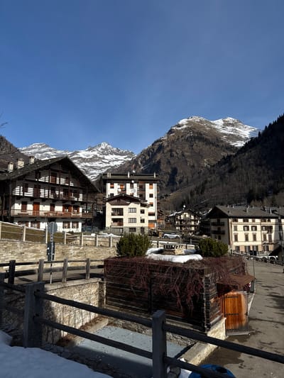 The image presents a panoramic view of a mountain village or town nestled beneath towering, snow-capped peaks under a clear, bright blue sky. The scene is captured during daytime, with direct sunlight illuminating the landscape and casting distinct shadows, suggesting either morning or early afternoon.
In the foreground and mid-ground, a collection of buildings of varying architectural styles forms the core of the settlement. To the far right, a wooden railing runs along what appears to be a path or elevated area. Behind it, a rustic wooden structure, possibly a shed or covered walkway, is visible, partially overgrown with reddish-brown, bare thorny bushes. A patch of snow is visible on the ground in the immediate foreground, indicating winter or early spring.
Towards the center-right, a multi-story building with a light-colored facade and numerous windows stands prominently. Further left, another multi-story building with a similar light-colored exterior features several satellite dishes on its roof or upper exterior walls, and some windows have dark shutters. To its left, a larger, more traditional-looking building with dark wooden walls and multiple stories, possibly featuring balconies or open-air corridors, dominates that section of the village. Scattered throughout the village are other buildings, some with light facades and dark roofs, blending into the mountainous backdrop. Stone walls and terracing are also visible, hinting at the challenging terrain.
The village is situated at the base of dramatic mountains. In the background, rugged, dark mountains rise majestically, with significant patches of snow clinging to their higher elevations, particularly on the prominent peak in the upper right. Further to the left, additional snow-capped peaks are visible against the clear sky. The lower slopes of the mountains are covered with a dense growth of dark, bare trees, transitioning into the built environment of the village. The bright blue sky fills the upper portions of the image, accentuating the clarity of the day.
No specific activity or individuals are visible, rendering the scene calm and tranquil. There is no visible text in the image.