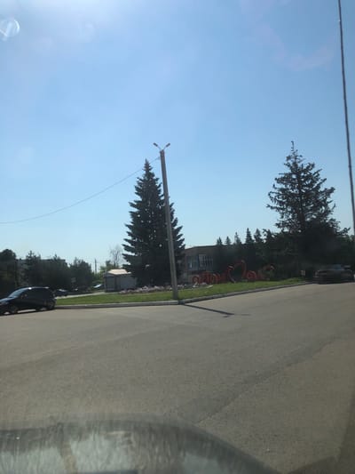 In Maksimovo, Russia, on a clear, sunny day, a street view shows a large Christmas tree in the foreground with a lamppost nearby. In the background, red decorative letters spelling "MAKSIMOVO" with a heart are visible in front of a building surrounded by trees. A dark car is parked on the street to the right of the letters, and another dark car is driving on the left side of the road.