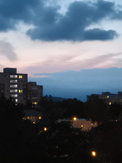 The image depicts a twilight scene in the city of Most, Czechia. The sky is filled with a mix of dark blue and light pink clouds. In the mid-ground, several apartment buildings are visible. The building on the left is taller and has many windows lit with a yellowish light, suggesting occupancy. To its right, another apartment building is partially obscured by trees, also showing some lit windows. The foreground is dominated by dark silhouettes of trees, with some scattered lights from the city visible through the foliage. The overall impression is of a residential area at dusk.