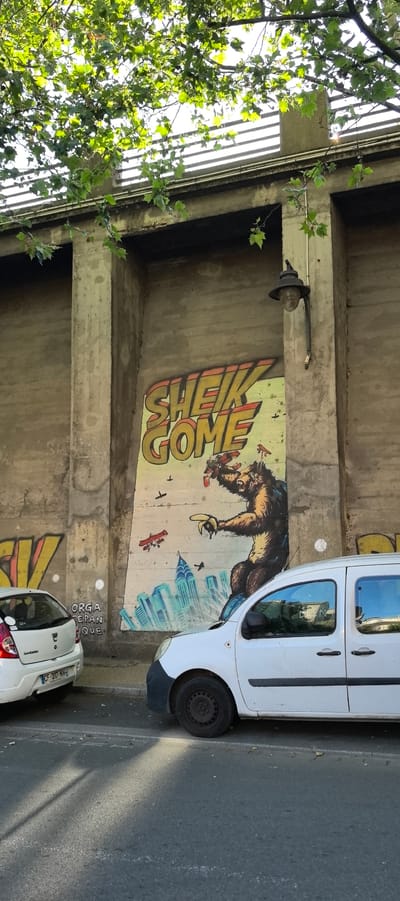 The image shows a section of a concrete wall in Villeneuve-Saint-Georges, France, featuring graffiti. On the wall, there is a large graffiti depicting a stylized ape or monster figure towering over a cityscape, with smaller airplanes flying around it. The text "SHEIK GOME" is prominently displayed in large, bold lettering above the figure. To the left of this main graffiti, there is another, less defined graffiti in yellow. In the foreground, two white vehicles are parked on the street: a smaller hatchback on the left and a van on the right. The van is partially obscuring the main graffiti. The scene is framed by the branches of a leafy tree overhead. The overall environment appears to be an urban setting, likely along a street or under an overpass, given the concrete structure and parked vehicles.