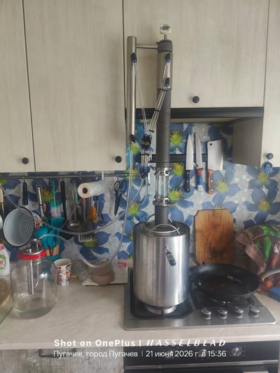 The image depicts a kitchen counter in Pugachyov, Russia, on June 21, 2026, at 3:36 PM. The focal point is a large distillation apparatus set up on a gas stove. The apparatus consists of a stainless steel pot, a thick insulated tube, and several clear plastic tubes connecting to a temperature gauge. Various kitchen utensils, including knives, a grater, brushes, and a roll of plastic wrap, are hung on a rack above the counter. A glass jar filled with liquid is also present, along with a mug and a frying pan on the stove. The background features light-colored cabinets and a floral-patterned backsplash. The scene suggests an ongoing distillation process.