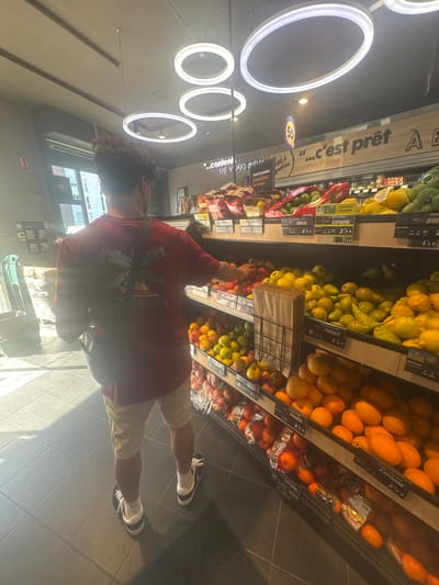 Here's a detailed description of the image:
**Scene:** The image captures a scene inside a supermarket, presumably in Marcq-en-Barœul, France, based on OCR data. The location appears to be the produce section.
**People:** A young man is standing in front of a display of fruits. He is wearing a red t-shirt with a design on the front, beige shorts, white socks, and black and white sneakers. He has dark curly hair. He is reaching towards the fruit display.
**Objects:**
* Displays of fruits (possibly oranges, lemons, limes, tangerines, and tomatoes) with price tags and labels indicating the origin of the produce (France, Spain, and Brazil).
* Supermarket shelving.
* Fluorescent ring lighting on the ceiling.
* A metal shopping cart in the lower part of the picture.
* Price tags and labels in French.
**Actions & Interactions:** The man appears to be selecting fruits from the display, possibly choosing which ones to purchase.
**Environment:** The store seems well-lit with tiled floors. There is a window in the background that indicates a building or street outside. The aisle is relatively clean.