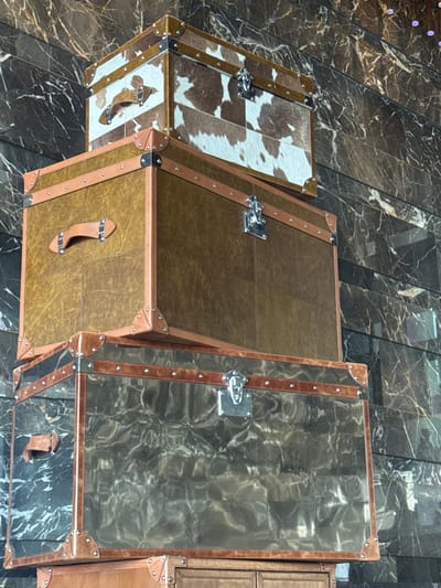 The scene is set in Bawshar Province, Oman, within an interior space against a dark, polished wall exhibiting distinct white and brown veining, consistent with marble, arranged in horizontal panels. Three distinct, vintage-style trunks are stacked vertically. No individuals are present.
The uppermost trunk is covered in brown and white cowhide patches, forming a mosaic pattern. It features light brown leather straps with silver rivets and corner reinforcements, a silver-toned metal latch on the front, and a brown leather handle on its left side.
Positioned beneath, the middle trunk is larger and constructed with a mottled, dark brown leather or faux leather material. It also has light brown leather straps, silver rivets, corner protectors, a silver-toned metal latch on the front, and a brown leather handle on its left side.
The lowest visible trunk features a dark, reflective surface with a wavy, textured pattern, possibly dark glass or a similarly reflective material. It is accented with light brown leather straps, silver rivets, corner protectors, and a silver-toned metal latch on its front. A brown leather handle is visible on its left side. The trunks are stationary, arranged in a stack against the detailed wall.