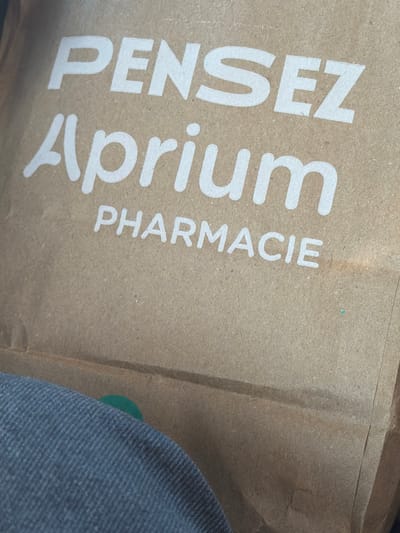 The image shows a close-up of a brown paper shopping bag. White text on the bag reads "PENSEZ Aprium PHARMACIE". The bag is made of recycled material and has a slightly textured surface. The bottom left of the image shows a portion of dark blue denim fabric, possibly clothing. A small, out-of-focus green circle is visible on the bag, near the denim. The lighting suggests an indoor environment, possibly a store or a home. The location is not visible or inferable from the image.