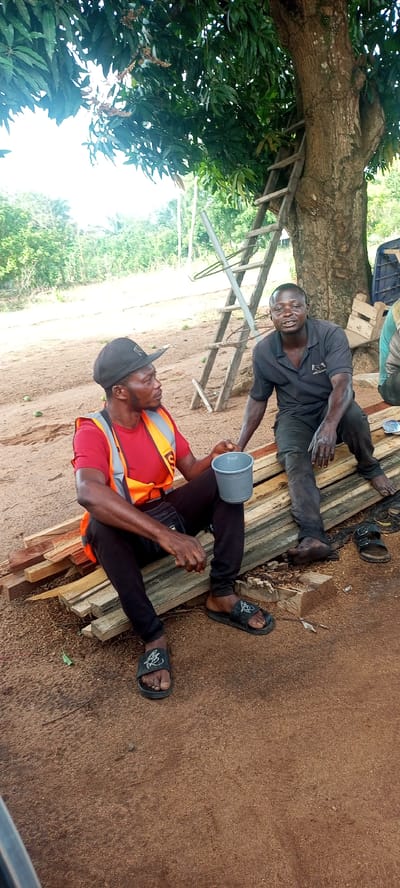 The image depicts two men sitting outdoors on a stack of wooden planks, engaged in conversation under the shade of a large tree in Iyara, Nigeria. The ground is reddish-brown and dusty.
The man on the left wears a black baseball cap, a red t-shirt, an orange safety vest with a visible "S" on it, black pants, and black slides. He holds a small, grey plastic bucket. The man on the right is dressed in a dark polo shirt with a faint logo and dark pants; his hands and bare feet appear soiled, indicative of manual work. His left sandal is on the ground beside him. He has an open, smiling expression, looking towards the man on the left.
In the background, a rustic wooden ladder leans against the trunk of the large tree. Beyond the tree, there's a expanse of green vegetation. The scene takes place during the daytime under natural light, suggesting a break from work in a rural or semi-rural setting, possibly involving timber or construction.