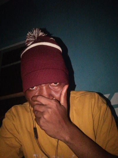 In Ajasse Ipo, Nigeria, a close-up, low-angle shot captures a person wearing a maroon beanie with a white pom-pom and stripe. The individual's face is partially obscured by their hand, which is covering their mouth. Their eyes are visible, with a slight redness in the conjunctiva. They are wearing a yellow t-shirt, and a black earbud wire hangs down from their neck. The background is a dark, textured wall, with some horizontal lines visible on the left side, possibly indicating a window frame or shelving. The lighting is dim, creating shadows that add to the overall mood of the image.