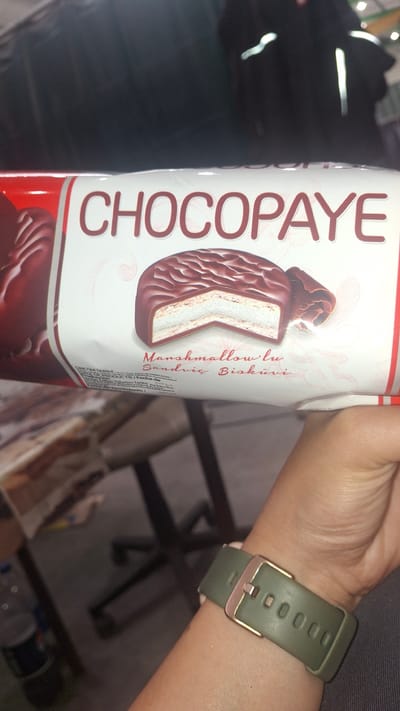 The image shows a close-up of a package of "CHOCOPAYE" cookies, which are described as "Marshmallow Sandwich Biscuits" in Turkish. The package features a cutaway view of one of the cookies, revealing its chocolate coating and marshmallow filling. A person's wrist, wearing a green watch with a rose gold buckle, is visible in the foreground, partially obscuring the lower portion of the packaging. The background is blurred, but appears to be an indoor setting with furniture. The location is stated to be Račice, Czechia.