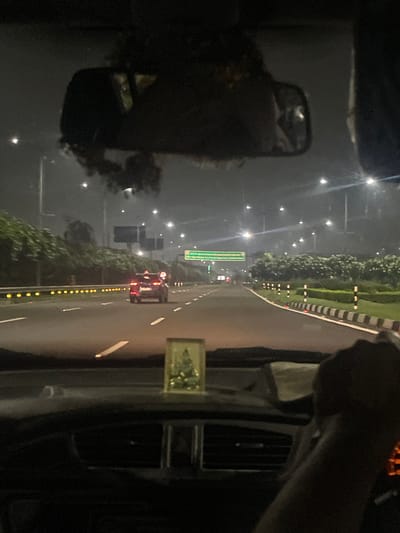 The image shows a nighttime view from inside a car driving on a multi-lane highway in Jhiurheri, India. The car's dashboard is visible in the foreground, with a small religious figurine placed on it. A hand is visible on the right side of the frame, suggesting the image was taken by a passenger.

The highway is illuminated by streetlights and the headlights of other vehicles. A dark-colored SUV is visible in the distance on the right lane. Green highway signs are present on the left side of the road. The road is lined with greenery and guardrails with reflective markers. The overall ambiance is dark and hazy, typical of nighttime highway driving in a city.