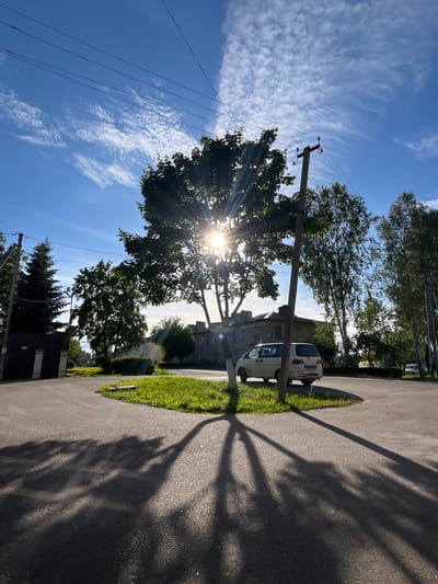 The image depicts a street scene in an urban or suburban environment in Serebryansky, Russia, under a bright, sunny sky with scattered wispy clouds. The sun is shining intensely, creating lens flare through a large tree in the foreground. The tree, with its green foliage, dominates the left side of the frame, casting a long, intricate shadow across the asphalt road on the right. A wooden utility pole stands near the tree, with power lines stretching across the sky.
A light-colored van is parked on the side of the road, next to a strip of green grass. In the background, a multi-story building with windows is visible. The overall scene suggests a warm, clear day, likely late afternoon given the long shadows. The environment appears residential or a quiet commercial area. There is no visible text in the image.