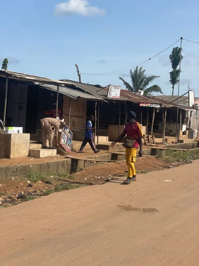 The image depicts a street scene in Ikoga, Nigeria. The street is unpaved, with buildings lining one side. The buildings appear to be constructed from corrugated metal and wood. Three people are visible: one is standing on the sidewalk, wearing a red shirt, yellow pants, a black cap, and a brown bag over his shoulder; another is walking nearby and wearing a blue shirt and black pants; another is sitting on the steps of the buildings, holding something. The sky is blue with scattered clouds. A palm tree and an antenna are visible. A poster is displayed on the front of the building.