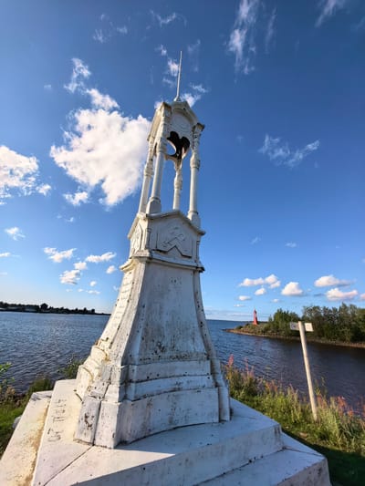 A white monument, possibly a bell tower, stands prominently in the foreground on a grassy bank next to a body of water. The monument is weathered and shows signs of age and wear. It has a tiered base and an open upper structure with pillars supporting a roof. A bell is visible inside the upper section. In the background, across the water, a red lighthouse stands on a small island or peninsula, surrounded by trees. The sky is blue with scattered white clouds, and the sun appears to be shining. The water is calm and reflects the sky. A white signpost with a white sign is visible to the right of the monument. The scene appears to be a public space, possibly a park or waterfront area in Nemyatovo-2, Russia, given the architectural style and the presence of a lighthouse.