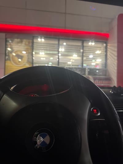 A close-up photograph shows the steering wheel of a BMW car. The steering wheel is black and features the BMW logo in the center. The background reveals a building with a red neon light outlining its edge and windows with illuminated displays. The blurred lights suggest an urban environment, potentially a store or business establishment. While the location is noted as Vodenichane, Bulgaria, specific contextual details beyond what is visible in the image cannot be determined.
