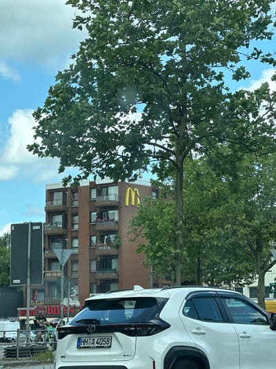 The image shows an urban scene in Seevetal, Germany. A white car is parked on the street. A building with a McDonald's logo on its facade is visible in the background. A Subway restaurant sign is also visible. There are trees with green leaves in the foreground, partially obscuring the building and the sky. The sky is blue with some clouds. A street sign is near the bottom of the frame. People are partially obscured behind the Subway sign. The license plate on the car reads "HH A 4058".