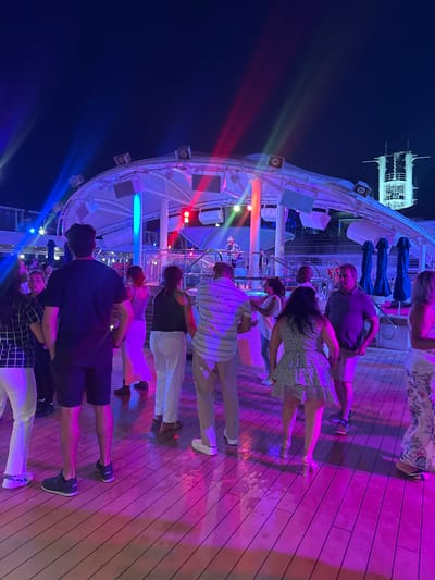 An outdoor nighttime party scene unfolds with a DJ on a raised platform illuminated by colorful stage lights. A crowd of people, dressed in casual attire, are dancing and socializing on a wooden deck. The background reveals a structured building, possibly a ship or a resort, with multiple levels and illuminated windows. The overall atmosphere is lively and festive, with vibrant lighting casting shadows and reflections across the scene. The location is not Salobreña, Spain; it appears to be a cruise ship deck party.