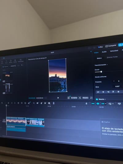 The image shows a close-up of a computer screen displaying video editing software, likely CapCut, given the visible "CapCut" logo. The software interface is predominantly dark, with various editing tools and timelines visible. In the main viewing window, a vertical video clip is displayed, showing a city skyline at sunset. The sky is a gradient of orange, pink, and purple, with buildings and a prominent tower visible in the distance. The video appears to be a landscape shot edited to fit a vertical format.

The bottom portion of the screen shows a timeline with multiple video tracks. Several video clips are loaded onto the timeline, indicated by file names like "HOOK2-090626-3.mp4" and "HOOK1-090626-1.mp4". These clips appear to contain footage of people and urban scenes.

On the right side of the screen, a panel displays editing options for the selected video clip, including "Transformación" (Transformation), "Escala" (Scale), "Posición" (Position), and "Girar" (Rotate). There is also an option for "Mezcla" (Blend).

A small pop-up window in the bottom right corner displays a "CapCut" logo and text in Spanish, stating "El atajo de teclado con otro existente" and "Grabar la pantalla: [Ctrl+A]". This translates to "Keyboard shortcut with another existing" and "Record screen: [Ctrl+A]".

The image does not contain any specific visual elements that would definitively place the scene in San Luis, Peru. The city skyline shown in the video clip is not identifiable as belonging to that specific location. The image primarily depicts a user engaged in video editing on a laptop.