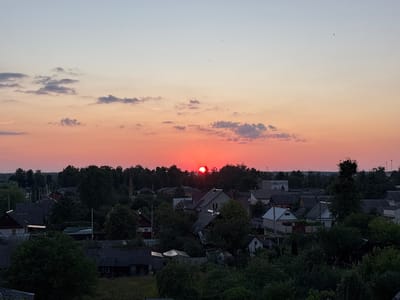 An aerial, horizontal, wide-angle shot captures a sunset over the village of Protasevichi, located within the Protasevichi Rural Council of Belarus. The sky is painted with hues of orange and pink, transitioning to a pale blue at the top. Silhouetted against the vibrant sky is a dense line of trees forming the horizon. Below the tree line, a cluster of houses with dark roofs and muted facades are visible, interspersed with more trees and greenery. The overall impression is that of a rural village at dusk.