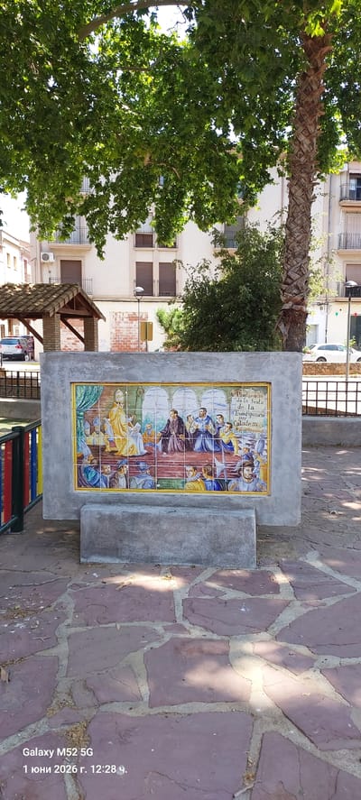 Here is a detailed description of the image:

The image shows a tiled monument in what is presumed to be a public area, likely a park or square, in the city of Canals, Spain. The monument features a large tile mural depicting a historical scene, possibly related to the "Proclamacio de la festa de la Transfiguracio per Calixte III". The mural shows figures in historical attire.

The monument is made of concrete with a rough, weathered texture. It is situated on a stone-paved surface. Various plants, including trees, shrubs, and a palm tree, are visible in the background and to the sides of the monument. Parts of a building are also visible behind the monument. A metal fence and what appears to be a set of colorful benches are also partially visible.

There is some text on the image indicating it was taken with a Galaxy M52 5G phone on June 1, 2026, at 12:28.