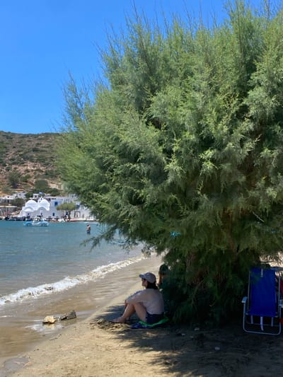 The image displays a sunny coastal scene within a bay or harbor area in the city of Apollonia, Greece. The foreground features a sandy beach with gentle waves breaking along the left edge. A large, dense green tree, possibly a tamarisk, occupies the right half of the frame, casting significant shade on the sand directly beneath it.
In the shaded area of the beach, a person, appearing female, is seated on the sand in the lower-center, facing left towards the water. She wears a light-colored long-sleeved top, dark shorts, and a light-colored baseball cap, with a blue and green mat visible beneath her. A second individual is partially obscured by the tree foliage behind the first person. On the far right, a blue folding beach chair is partially visible under the tree.
The middle ground consists of calm, blue-green water. One person is standing chest-deep in the water, facing away from the camera towards the distant shore. A light-blue, car-shaped paddle boat is moored in the water further out. The background presents a cluster of white Cycladic-style buildings with characteristic rounded roofs and domes, forming a village or town along the distant coastline. Several smaller boats are docked near these structures. Behind the village, a dry, rocky hillside rises under a clear, bright blue sky, indicating fair weather.