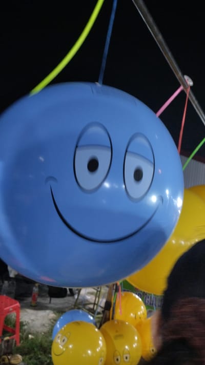 A large, light blue balloon with a cartoon face, featuring wide, black-pupiled eyes and a wide smiling mouth, dominates the foreground of this outdoor image. The balloon is suspended by colorful strings, including lime green, blue, and pink, which are attached to a metal pole overhead. The scene appears to be an outdoor fair or event, likely at night or dusk, given the dark background and the bright artificial lighting reflecting off the balloons.
Behind the main blue balloon, several yellow balloons are visible, some also decorated with cartoon faces, suggesting a festive atmosphere. One yellow balloon in the lower right has a furry, dark object partially obscuring it, possibly a person's clothing or hair. In the lower left, a red stool and some greenery can be seen, indicating a ground-level setting. The overall impression is one of a cheerful and playful gathering. No visible text is present in the image. The location context of Sambungrejo, Indonesia, suggests this could be a local celebration or market.