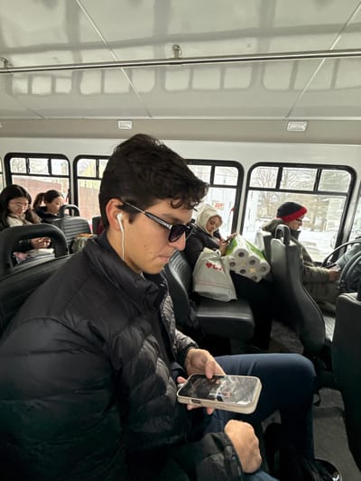 Inside a bus with white interiors and multiple windows, passengers are seated. A man with dark hair, sunglasses, and an earbud is in the foreground, looking at his phone. Behind him, a woman with a white headscarf sits next to a stack of toilet paper and a shopping bag. The driver is visible in the distance, wearing a red and black beanie. Several other passengers are also present, and the outside scenery is visible through the windows.