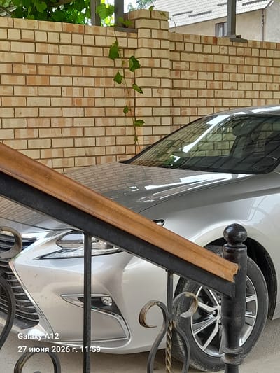 A silver sedan is parked outdoors, with its front section partially visible. The car is positioned in front of a light brown brick wall. A dark metal railing with a wooden handrail runs diagonally across the foreground, obscuring a portion of the vehicle. The car's headlights and grille are visible, as is its front tire and wheel. Text overlay on the bottom left corner indicates the device used is a "Galaxy A12" and the date and time are "27 июня 2026 г. 11:59". The environment appears to be a residential or commercial outdoor area with some vegetation visible in the upper left corner. The scene is likely in Kara-Köl City, Kyrgyzstan, based on the date format and Cyrillic text.