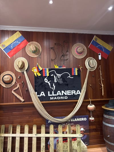 The image depicts the interior of a Venezuelan restaurant, "La Llanera," located in Torrejón de Ardoz, Spain. The walls are adorned with Venezuelan flags, traditional straw hats, and decorative items like a hammock, a small harp, and various trinkets. A prominent black banner displays the restaurant's name and location, "LA LLANERA MADRID," along with a stylized outline of a cow. In the foreground, a rustic wooden fence encloses a display of hay, and a poster for "La Llanera Edicion" features a man in a cowboy hat. To the right, a large wooden barrel and a traditional Venezuelan hat contribute to the cultural ambiance. The lighting suggests an indoor setting, possibly a dining area or a themed section of the restaurant.
