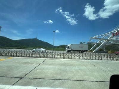 The image captures a daytime scene on a highway in Hoa Xuan, Vietnam, under a bright blue sky with scattered white clouds. The foreground shows a concrete road with distinct lane markings. A metal accordion-style gate system is positioned along the side of the road, suggesting a checkpoint or toll booth area.
In the middle ground, a white sedan is driving on the highway, and to its right, a white truck with a cargo container is also in motion. The truck displays text that appears to be in Vietnamese. Visible on the truck is a phone number: "0918.602.402" and "0912.202.408", and "63A-582.02". In the background, lush green mountains rise, with one prominent peak in the distance. A large, modern, white metal structure, possibly part of a bridge or overpass, is visible on the right side of the image, featuring red lettering that appears to be the word "TR" followed by other letters. The overall scene conveys a sense of travel and transit on a clear day.