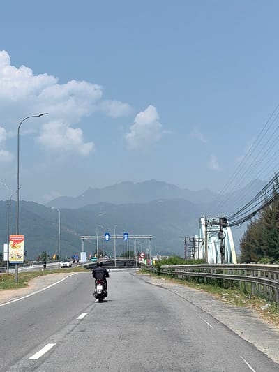The image captures a highway scene in Hải Vân Ward, Vietnam, under a clear blue sky with scattered white clouds. A paved road, marked with white solid and broken lines, extends into the distance. A single individual, wearing a dark jacket, operates a white scooter in the center of the road, moving away from the viewer. To the right, a metal guardrail parallels the road, beyond which lies overgrown vegetation and pine trees. On the left, multiple streetlights line the roadside, along with a tall pole displaying a vertical, colorful billboard advertisement. Further down the road, a white car and other vehicles are visible approaching an overhead gantry structure with blue informational signs. To the right, a light blue metal truss bridge is prominent, with numerous black power lines and cables extending across the upper right portion of the frame and attached to structures near the bridge. A range of hazy, blue-grey mountains forms the background, spanning the entire horizon.