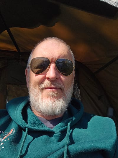 A close-up, rotated image shows an older man with a bald head and white beard wearing gold-rimmed sunglasses. The man is wearing a dark green hooded sweatshirt. The background appears to be the interior of a tent or shelter, with the tent walls in a brown color. The lighting suggests an outdoor environment with direct sunlight. The location, Gavisse, France, cannot be confirmed as there are no identifiable landmarks or location markers in the image.