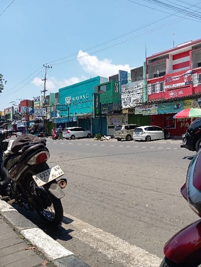The image depicts a daytime street scene in Pasuruan, Indonesia, characterized by commercial activity along a main thoroughfare. In the immediate foreground on the left, the rear section of a dark-colored motorcycle, equipped with a top bag on its seat and an Indonesian license plate "H 6927 BH" (expiration 04-28), is parked on a sidewalk curb. On the far right foreground, a portion of a red/maroon scooter's front fender and wheel is visible.
The midground features an asphalt road with visible white dashed and solid lane markings. Several vehicles are present: two white multi-purpose vehicles (MPVs) are parked consecutively on the right side of the road, followed by a beige MPV. A white sedan or smaller vehicle is visible further down the street on the left. Scattered individuals are present on the sidewalks near the storefronts and vehicles, including at least one person in a red top. A street vendor's stall, identified by a red, green, and yellow striped umbrella, is partially visible on the right side of the road.
The background is dominated by a row of multi-story commercial buildings lining the street. Prominent storefronts include a turquoise building labeled "MAYANG," a green building with "Pegadaian" written vertically, and a red and white building displaying "JOTUN PAINT" and "SAMBOS JAYA." Overhead electrical or communication wires crisscross the clear blue sky, which contains a few scattered white clouds. A utility pole stands on the left side of the street. The scene is well-lit, indicating a sunny day.