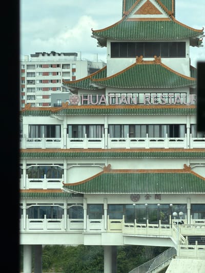 This is a photograph of a building with a distinctive East Asian architectural style. The building features ornate green tiled roofs with curved eaves and decorative elements. The facade is a combination of light-colored walls, white trim around windows and doorways, and a prominent pink neon sign with the text "HUA TIAN RESTAURANT" running vertically.
The building appears to be a commercial establishment, likely a restaurant. Several windows and doors are visible, some with intricate white framing. There are also some decorative architectural elements, such as what appear to be carved figures or symbols, adorning the building's exterior. To the right of the restaurant entrance, there's an outdoor staircase made of white metal railings. The background includes lush green foliage and a cloudy sky, suggesting an outdoor setting. The photograph is taken from an elevated perspective, looking down and across the building. The orientation of the image is rotated 90 degrees counter-clockwise. The specific location is Alfortville, France. No people are visible in the frame.