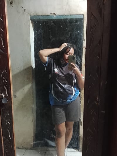 A young woman is taking a selfie in a mirror. She is wearing a black t-shirt with blue and white accents and gray shorts. Her right hand is on her head, and she is holding a smartphone in her left hand. The mirror is in a room with a textured wall and a wooden door frame with carved designs. The floor is tiled. The location is the city of Gunung Malang, Indonesia.