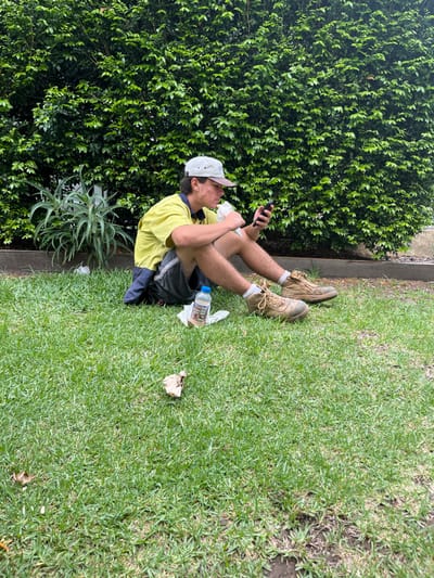 A man in a yellow work shirt, dark shorts, and a white cap sits on green grass near a dense bush. He wears tan work boots and white socks, and holds a phone in one hand and something wrapped in paper in the other. Next to him is a water bottle, discarded food packaging, and a black bag. The photo is rotated sideways.