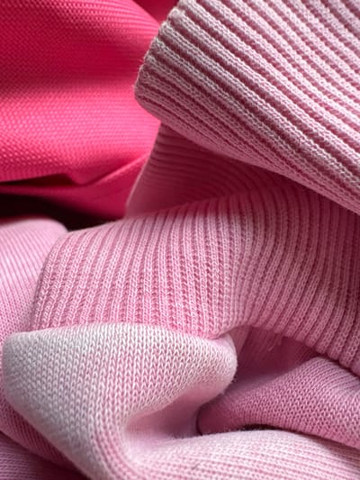 A close-up, overhead shot focuses on a pile of pink knit fabric. The fabric exhibits ribbed textures and varying shades of pink, with some areas appearing almost white. The lighting highlights the folds and contours of the material, creating a soft, textured visual. The scene is devoid of people or discernible environmental context, making it impossible to determine its location within Гарадзецкі сельскі Савет, Belarus.