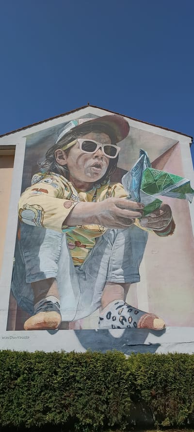The mural on the side of a building in Siero, Spain, depicts a young person wearing a baseball cap, sunglasses, and a yellow shirt with cartoon characters. The individual is crouched down and holding a crumpled green and white packet of snacks. The mural is painted on a building with a tiled roof, and below it is a dense green hedge. The sky is clear and blue. The mural is signed with the name "DADOSPUNTOCERO".