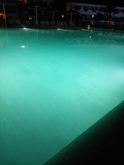The image depicts an outdoor swimming pool at night, illuminated by underwater lights which cast a vibrant turquoise glow. The water is calm with slight ripples. The edge of the pool, a dark, textured surface, occupies the lower right portion of the frame. In the background, a building with visible windows that are lit from within, along with several lounge chairs and umbrellas, suggest a hotel or resort setting. The presence of palm trees further indicates a warm climate. The overall scene is serene and tranquil, characteristic of a nighttime ambiance at a resort. The location is identified as Kos, Greece.