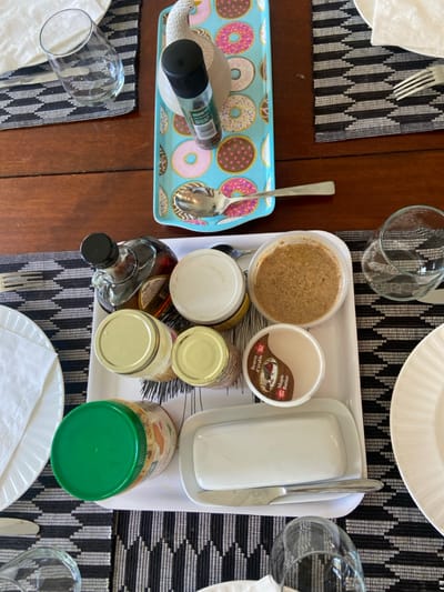 The image shows a table setting in Rye, New York (coordinates: 40.952, -73.708). There are multiple jars, containers, and a butter dish on a white serving tray. The jars contain various foods, including maple syrup, peanut butter, and other condiments. A spoon sits on top of a blue tray with a colorful donut pattern. Two glasses are present, and there are two white plates on the table, one with a fork next to it. The table is covered with a black and white checkered tablecloth. A white plate is on the table to the right of the tray. A white plate is on the table to the left of the tray. There are also two glasses of water on the table, one to the left of the tray and one to the right of the tray. The table setting appears to be prepared for a meal.