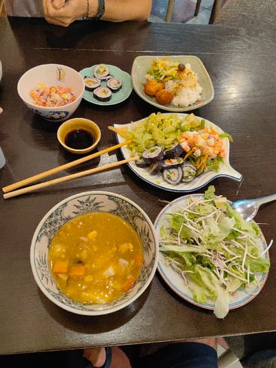 A top-down view shows a dark brown table laden with a meal for at least two individuals in a restaurant setting within Hòa Cường Ward, Vietnam. In the upper left, a white ceramic bowl with a colorful geometric pattern contains a salad of diced vegetables and possibly imitation crab meat. Adjacent to it, a small, light green, leaf-shaped plate holds six sushi rolls. To the upper right, a square, light green plate presents white rice, stir-fried vegetables, and two golden-brown fried croquettes. A small, light brown ceramic bowl contains dark soy sauce. Two pairs of light wooden chopsticks are positioned on the table; one pair rests partially over the soy sauce bowl, and the other extends across a plate. A large, white, leaf-shaped plate, centrally located on the right, holds a bitter melon stir-fry, a separate portion of a creamy salad with visible red and orange pieces, and several additional sushi rolls, garnished with a lime wedge. In the lower left, a white ceramic bowl with a blue and white patterned rim contains a yellow soup or curry with diced root vegetables and corn kernels. To its right, a round, white ceramic plate with a thin blue rim holds a fresh green salad of torn lettuce and bean sprouts, with a metal spoon resting upon it. The forearms and hands of one person, wearing a dark bracelet, are visible at the top-left edge of the table. The lower legs and feet of another individual, clad in dark trousers and light-colored flip-flops, are partially visible beneath the table at the bottom.