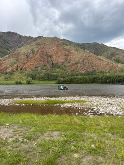 The image displays a river landscape in Burentogtoh, Mongolia, under an overcast sky. A light blue, small truck, with its bed loaded and covered by tarps, is positioned in the center of a wide, shallow river, suggesting a ford crossing. The river's immediate banks and parts of its bed are visible, composed of light-colored pebbles and rocks. Beyond the river, a range of mountains rises steeply, characterized by reddish-brown rock formations and slopes covered in green vegetation and scattered trees. A dense line of dark green trees runs along the far bank of the river at the base of the mountains. The foreground features a grassy riverbank with patches of exposed earth and small stones. No individuals are visible.