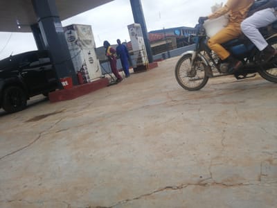 The image depicts a scene at a gas station in Ajasse Ipo, Nigeria. In the foreground, a black SUV is parked. To the right, a motorcycle carrying two individuals is moving away from the camera. The driver is wearing a yellow outfit, and the passenger is in white pants. The background shows gas pumps with "MRS" branding and two individuals, one in a blue uniform and another in a maroon and yellow uniform, standing near a pump. A red fire extinguisher is visible next to the SUV. The ground is a cracked concrete surface. The sky is overcast.