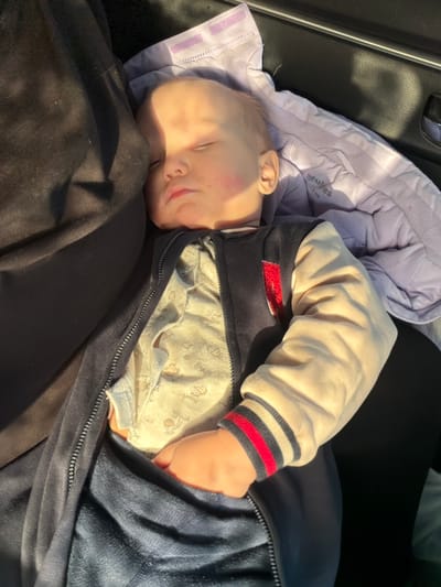 Within a vehicle in the city of Abdes-Urdes, Russia, an infant is depicted sleeping. The subject, a pale-skinned infant with light hair, is positioned with eyes closed and mouth slightly open. Both cheeks exhibit a faint reddish coloration. The infant wears a light-colored, patterned long-sleeved undershirt, covered by a navy blue zippered jacket featuring cream-colored sleeves with red, navy blue, and cream striped cuffs. The infant's right hand is tucked into the lower portion of the navy blue garment. Behind the infant's head and upper back is a light purple, padded fabric item, possibly a car seat insert, displaying "iules" and "car" text and velcro straps. A dark fabric object is partially visible to the infant's left. In the upper right background, a silver-colored car door handle is visible. Strong sunlight illuminates the infant's face and upper torso, casting pronounced highlights and shadows. The infant appears to be resting within a car seat or similar vehicle restraint system.