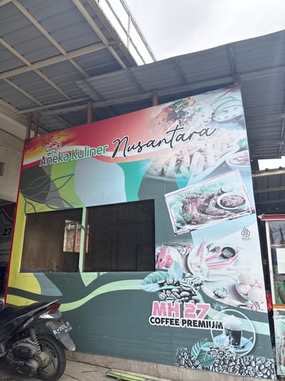 The image shows a large, outdoor advertisement banner displayed on the side of a building. The banner is primarily green and features images of food and coffee. Text on the banner includes "Aneka Kuliner Nusantara," "MH27 COFFEE PREMIUM," and "HALAL INDONESIA." There are also images of coffee beans, crab, eggs, and what appears to be a plate of food. The banner is partially obscured by the roof of a building, which appears to be made of corrugated metal. A motorcycle is parked in front of the banner. The setting suggests a street or an outdoor commercial area in Meunasahmanyet, Indonesia. The time of day is unclear, but the lighting suggests it could be daytime. There are no people visible in the image.