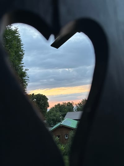 The image, taken in Voronov, Russia, is a framed view of a rural landscape through a heart-shaped opening in a dark foreground structure. The sky dominates the upper portion of the frame, filled with a gradient of soft gray clouds, transitioning to lighter hues with hints of sunset colors like yellow and pink near the horizon. Below the sky, a dense canopy of green trees stretches across the middle ground. Nestled amongst the trees are two buildings: a small, rustic wooden log cabin with a green roof and a single window, and a larger building with a darker, possibly gray tiled roof. The setting appears to be a tranquil evening.