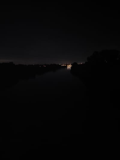 The image shows a nighttime scene of the Main River in Flörsheim, Germany. The foreground is dominated by darkness, obscuring the water and riverbanks. In the distance, on the far bank, a city skyline is visible with numerous illuminated buildings and lights reflecting on the water. The silhouette of trees is visible on the right side of the image, bordering the river. The overall impression is one of a quiet, urban nighttime landscape.