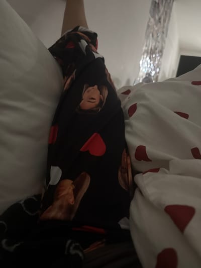 A close-up shot shows a person lying in bed, mostly covered by white bedding with red heart patterns. The person is wearing dark pajamas featuring images of a woman's face, accompanied by red and white hearts. Only a small portion of the person's arm is visible. The background reveals a glimpse of a room with a blurry, light-reflecting surface. The overall perspective is slightly angled downward, focusing on the bedding and pajamas.