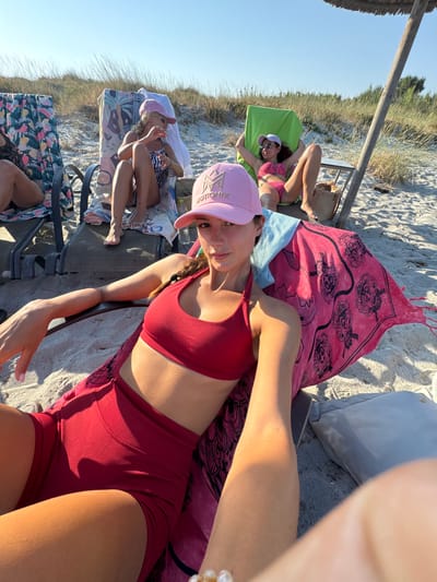 The image depicts a group of young women relaxing on a beach in Greece. The scene is set on a sandy beach with dry grass and vegetation visible in the background, suggesting a sunny and warm day. In the foreground, a woman wearing a red bikini and a pink baseball cap is lying on a beach chair, looking towards the camera. Another woman in a pink and black striped bikini is also visible lying on a chair to the left. Several beach chairs are arranged on the sand, some covered with colorful towels. A large pink towel with a black graphic pattern is spread out on one of the chairs. The time of day appears to be late afternoon or early evening, as indicated by the long shadows cast by the chairs and vegetation, and the warm, golden light. The overall mood is relaxed and casual, with the women enjoying a day at the beach. The baseball cap worn by the woman in the foreground has the word "MOTION" visible on it.