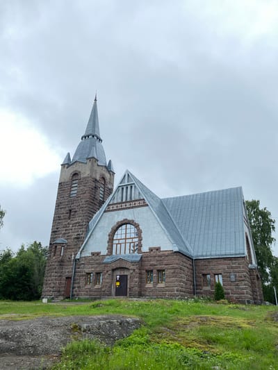 The image shows a stone church with a tall, pointed steeple, located in the city of Melnikovo, Russia. The church is constructed from rough-hewn stones, predominantly in shades of brown and grey. The roof of the main building and the steeple are made of metal, with a silvery-grey hue. The building features several windows, some with arched tops and ornate stonework surrounding them. There is a large arched window above the entrance. The church is situated on a grassy hill, with patches of moss and wildflowers visible. Large rocks are present in the foreground, adding to the natural landscape. Trees and foliage surround the church, with one large tree visible to the right of the building. The sky is overcast, with a grey, cloudy appearance. No people are visible in the image. A small plaque is mounted on the wall of the church near a small evergreen tree.