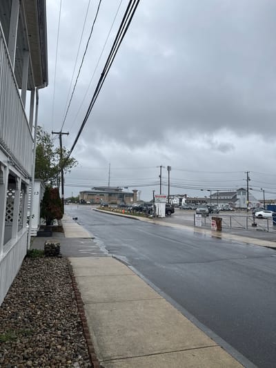 The image depicts a wet, overcast street scene in Hampton, Virginia. In the foreground, part of a white-painted building with a slatted awning or roof is visible. A narrow strip of gravel and small plants borders the building. A grey concrete sidewalk runs alongside a wet asphalt street. Power lines stretch across the sky.
Across the street, a parking lot with several parked cars and signage is seen. Beyond the parking lot, a mix of low-rise buildings and structures are visible. The general environment suggests a coastal or urban area. No people are visible in the image.